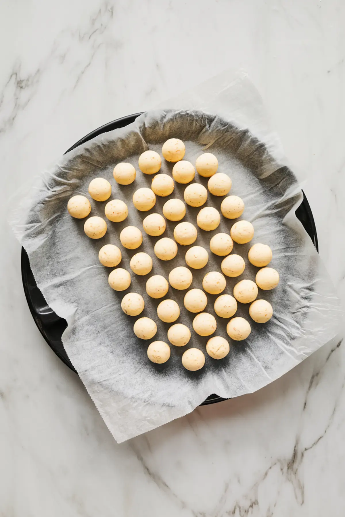 Tray of evenly spaced, raw cookie dough balls lined on parchment paper, ready for baking, arranged in a heart shape on a round black baking tray over a marble surface.
