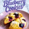 Close-up of baked lemon blueberry cookies with a golden crust and bursting blueberries, stacked on a pink plate and sprinkled with powdered sugar, with text overlay reading “Lemon Blueberry Cookies.”