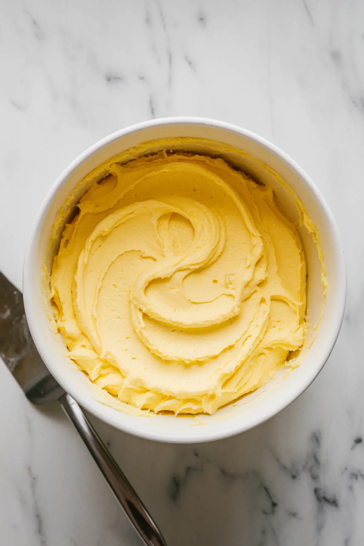 A white bowl filled with smooth lemon buttercream frosting swirled in a circular pattern on a marble countertop, with a metal spatula resting nearby.
