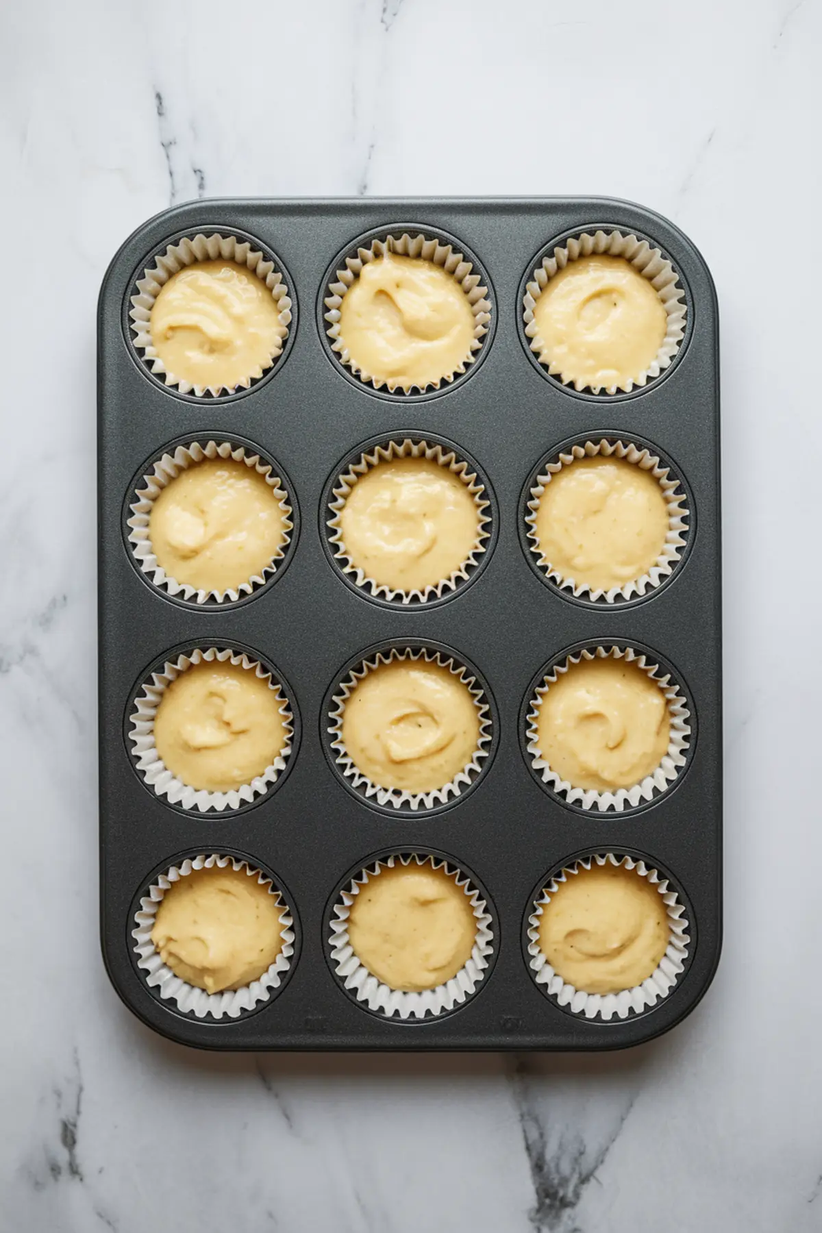 Cupcake tray filled with twelve unbaked lemon cupcake liners, each holding a scoop of smooth yellow batter, placed on a white marble countertop.
