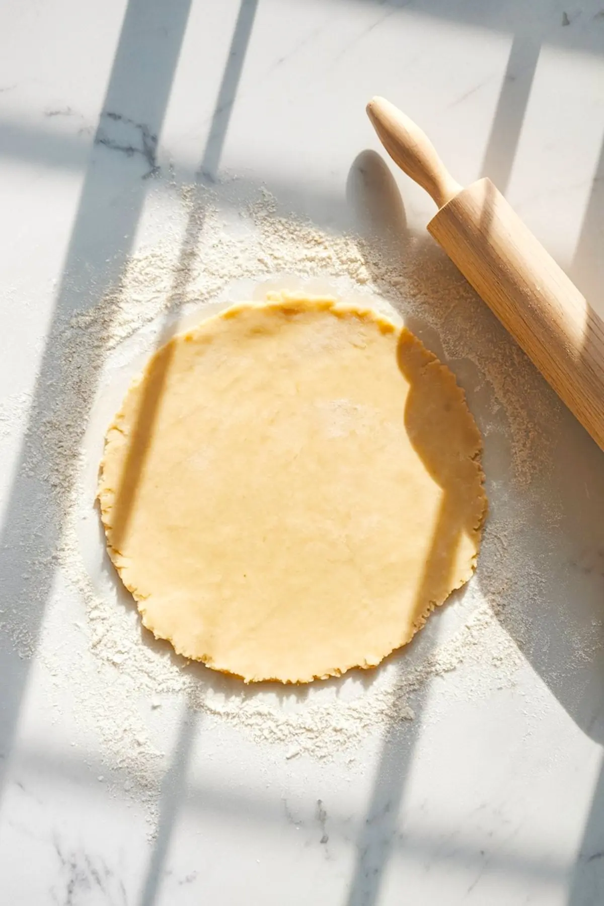 Round disc of pie dough rolled out on a floured marble surface, with a wooden rolling pin on the side and sunlight casting shadows.

