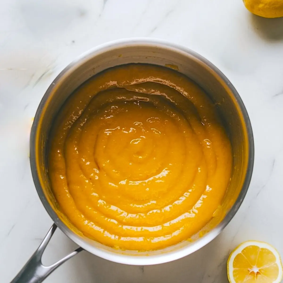 Saucepan containing thick, glossy yellow lemon curd, with a halved lemon placed next to it on a marble surface.
