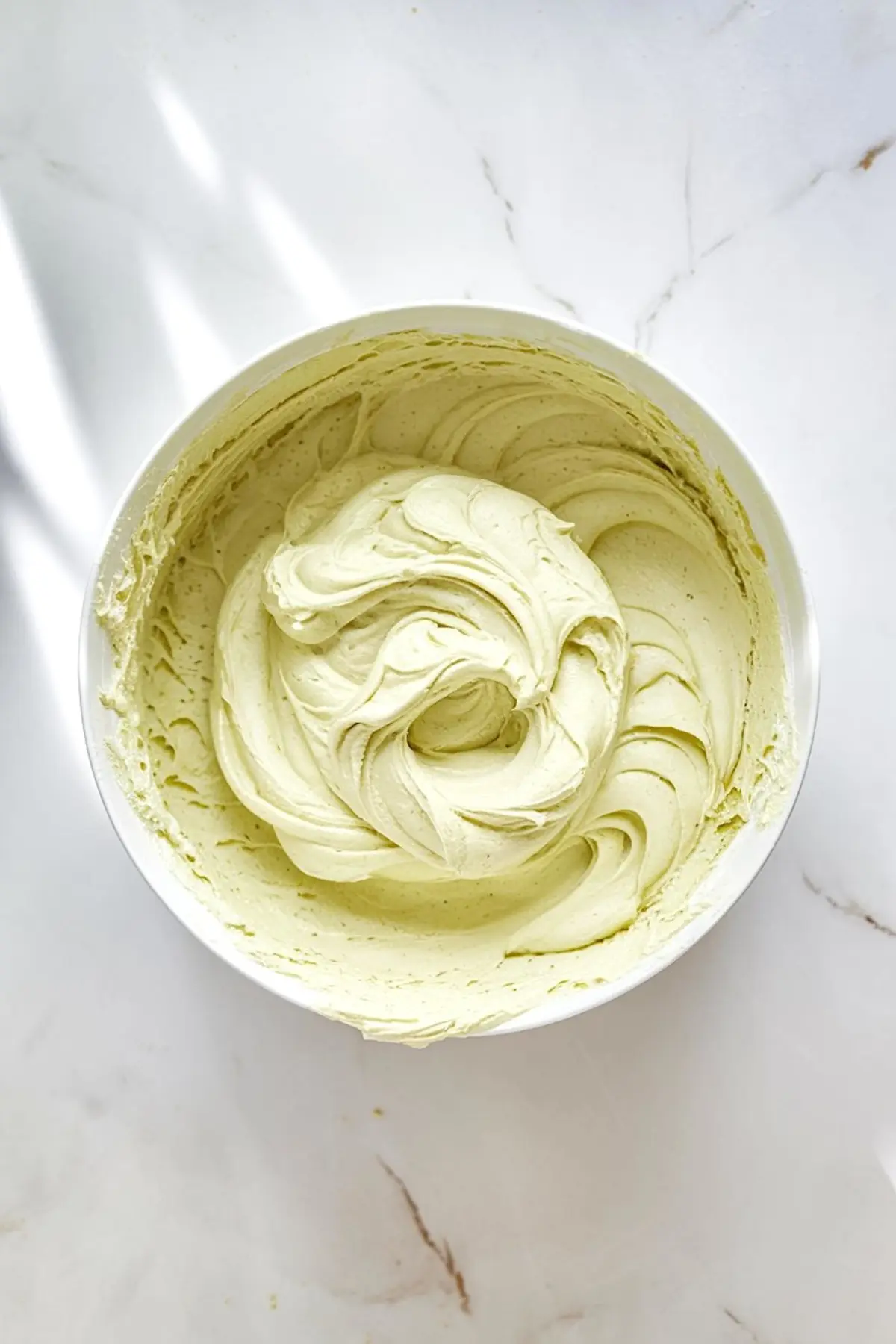 Close-up image of a mixing bowl filled with whipped pistachio buttercream frosting. The frosting is light green with smooth, creamy texture and soft swirls, sitting on a white marble surface.