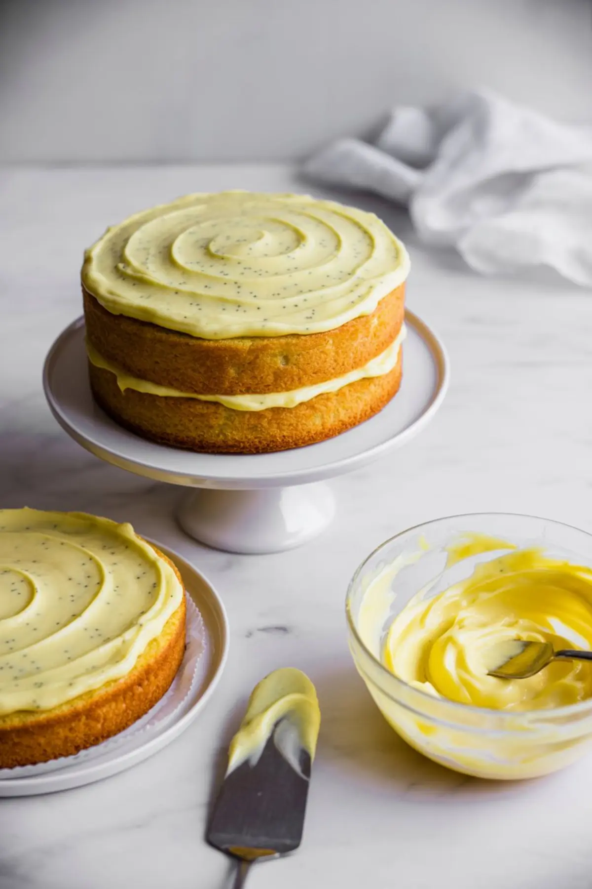 Tall layer cake with lemon poppy seed sponge and pale yellow frosting is being assembled on a white cake stand, with frosting swirled on top and a bowl of frosting nearby on a marble countertop.