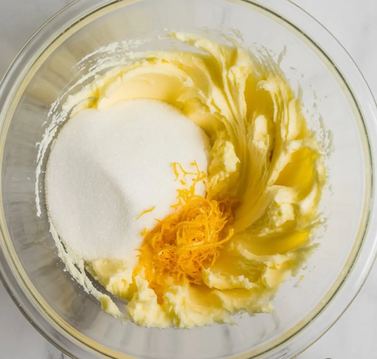 Butter and sugar mixture in a mixing bowl with fresh lemon zest, showing the creaming stage of cake preparation on a marble background.