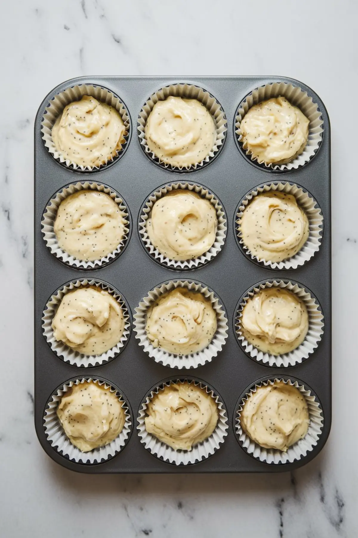 Muffin tray filled with raw lemon poppy seed batter in white paper liners, ready for baking.