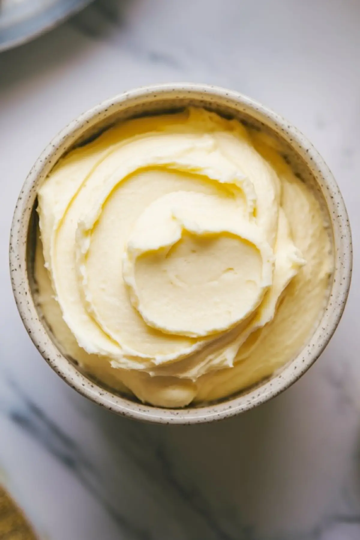 Overhead view of a ceramic bowl filled with smooth, swirled vanilla buttercream frosting on a marble surface.