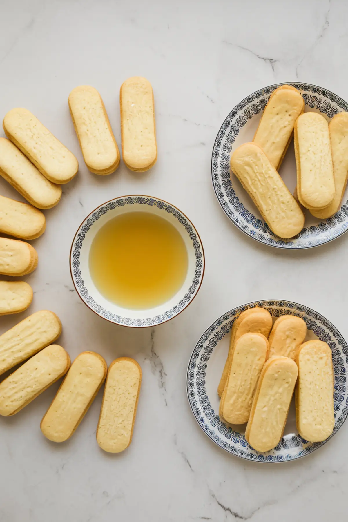 Flat lay of ladyfinger cookies arranged around a patterned bowl of lemon juice on a marble surface, with extra cookies served on decorative plates for a lemon tiramisu recipe.