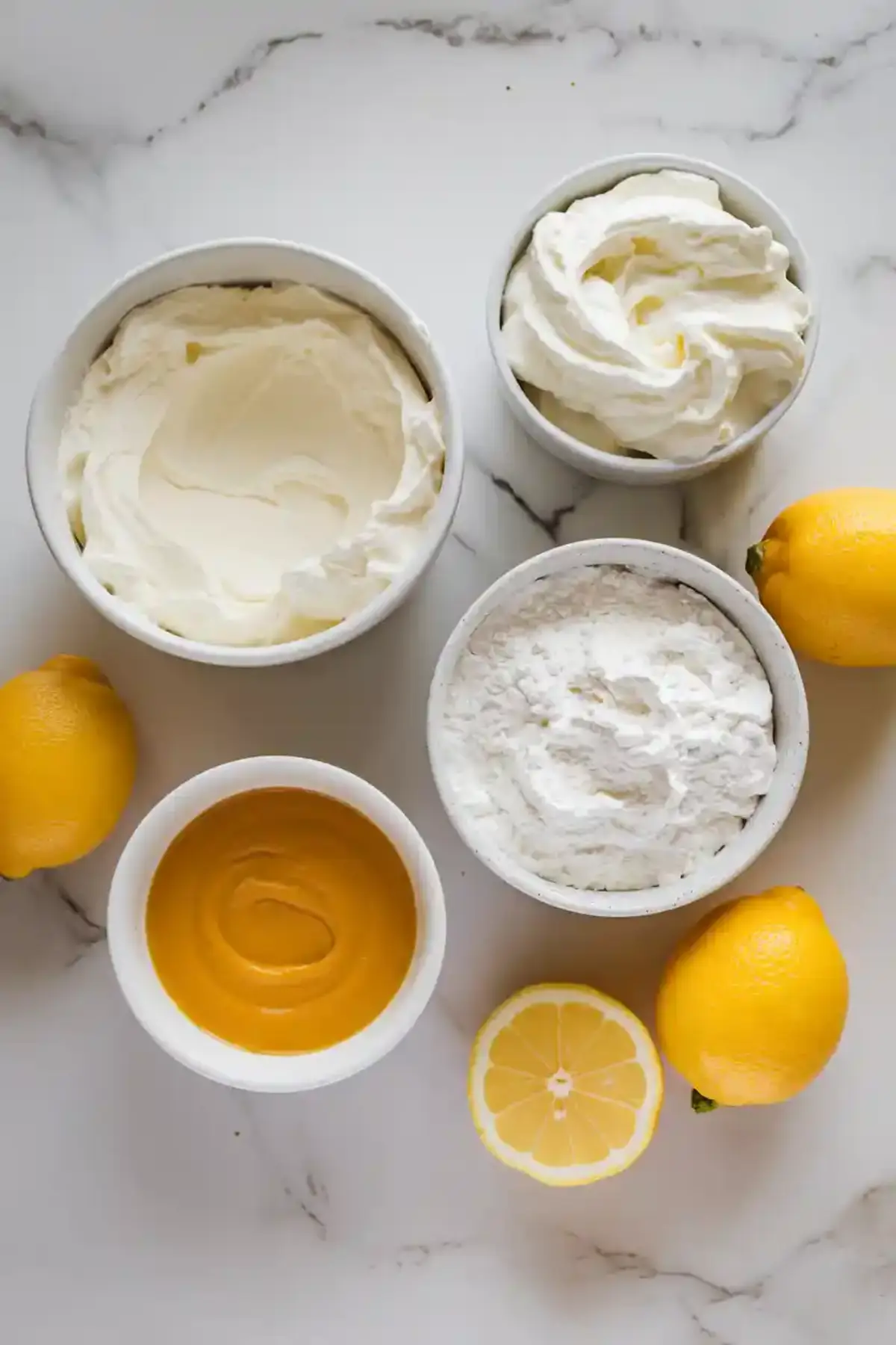 Overhead view of dessert ingredients including mascarpone, whipped cream, powdered sugar, lemon curd, and fresh lemons on a marble background, prepared for making lemon tiramisu.