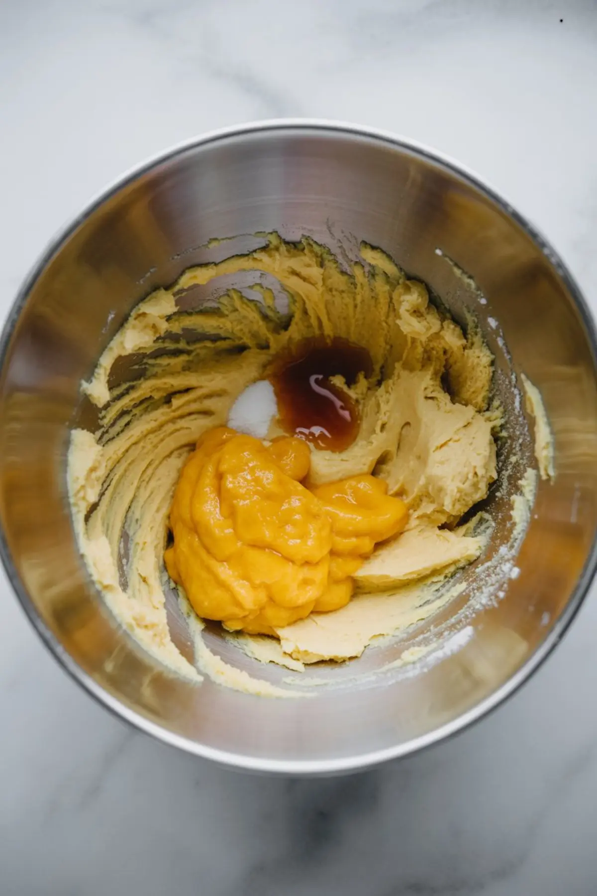 Metal mixing bowl with blended cookie dough and added mango puree, vanilla, and salt, resting on a marble surface ready for mixing.