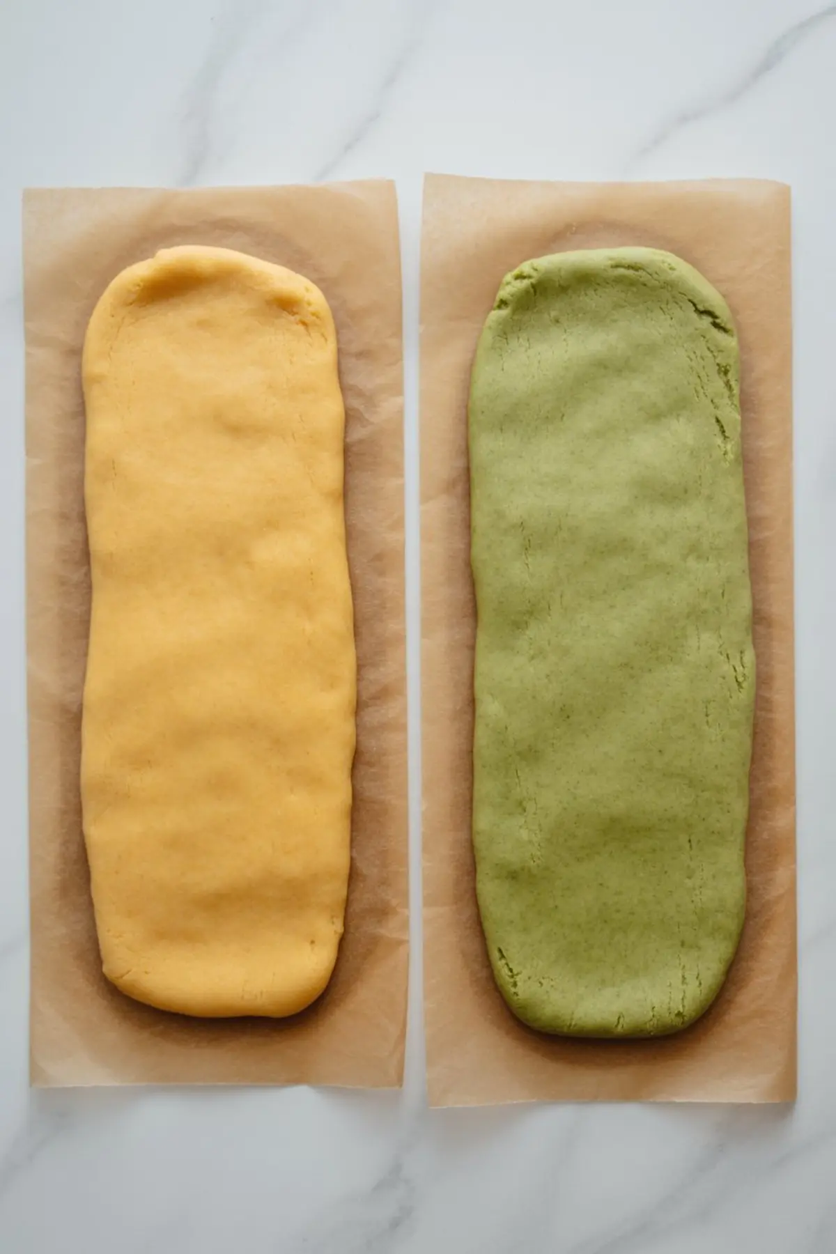 Two slabs of cookie dough on parchment paper, one bright yellow mango-flavored and one muted green matcha-flavored, placed side by side on a marble countertop.