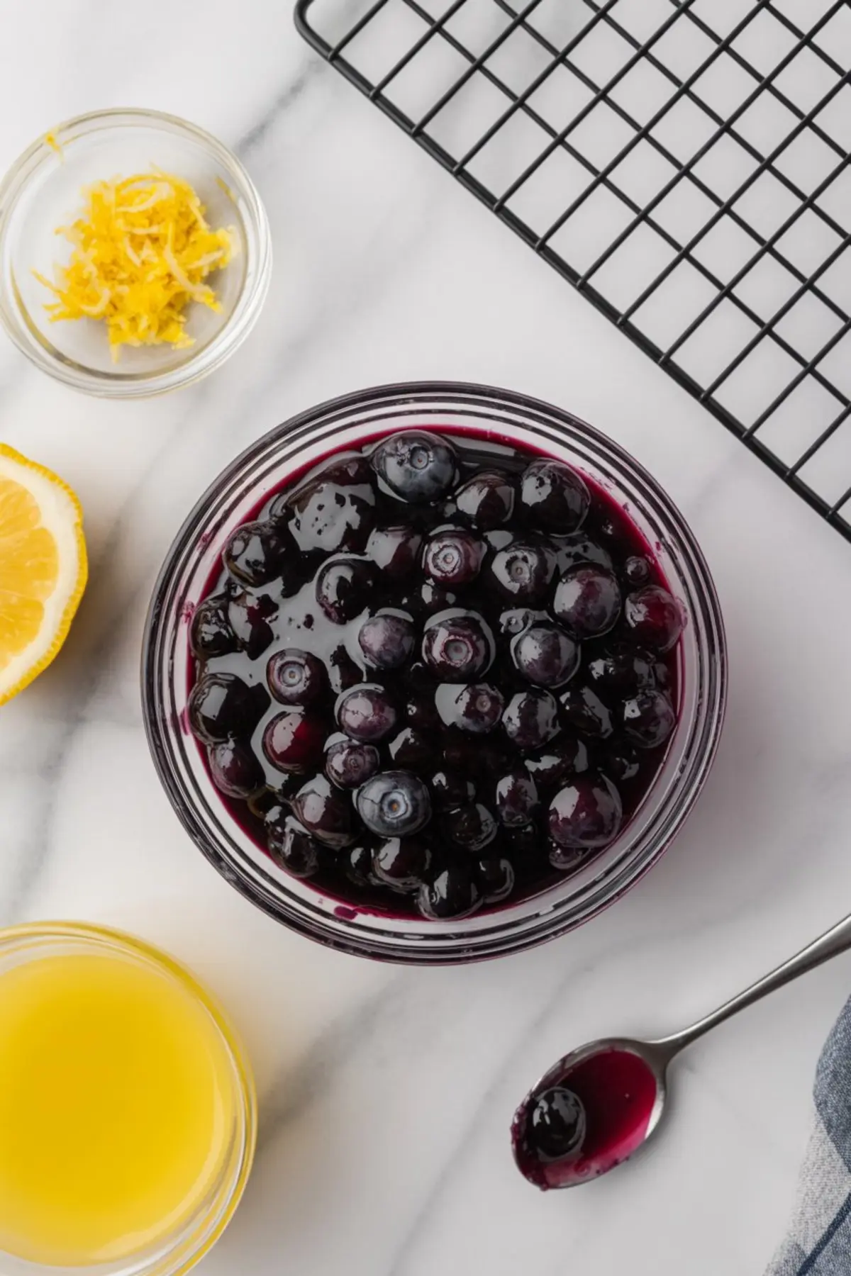 Glass bowl of glossy blueberry compote with whole blueberries, placed near lemon zest, lemon juice, and a cooling rack on a marble countertop.