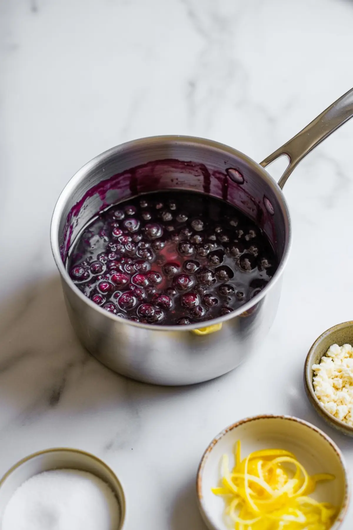 Saucepan filled with cooked blueberry filling on a marble countertop, surrounded by lemon zest, sugar, and crumbled cheese in small dishes.