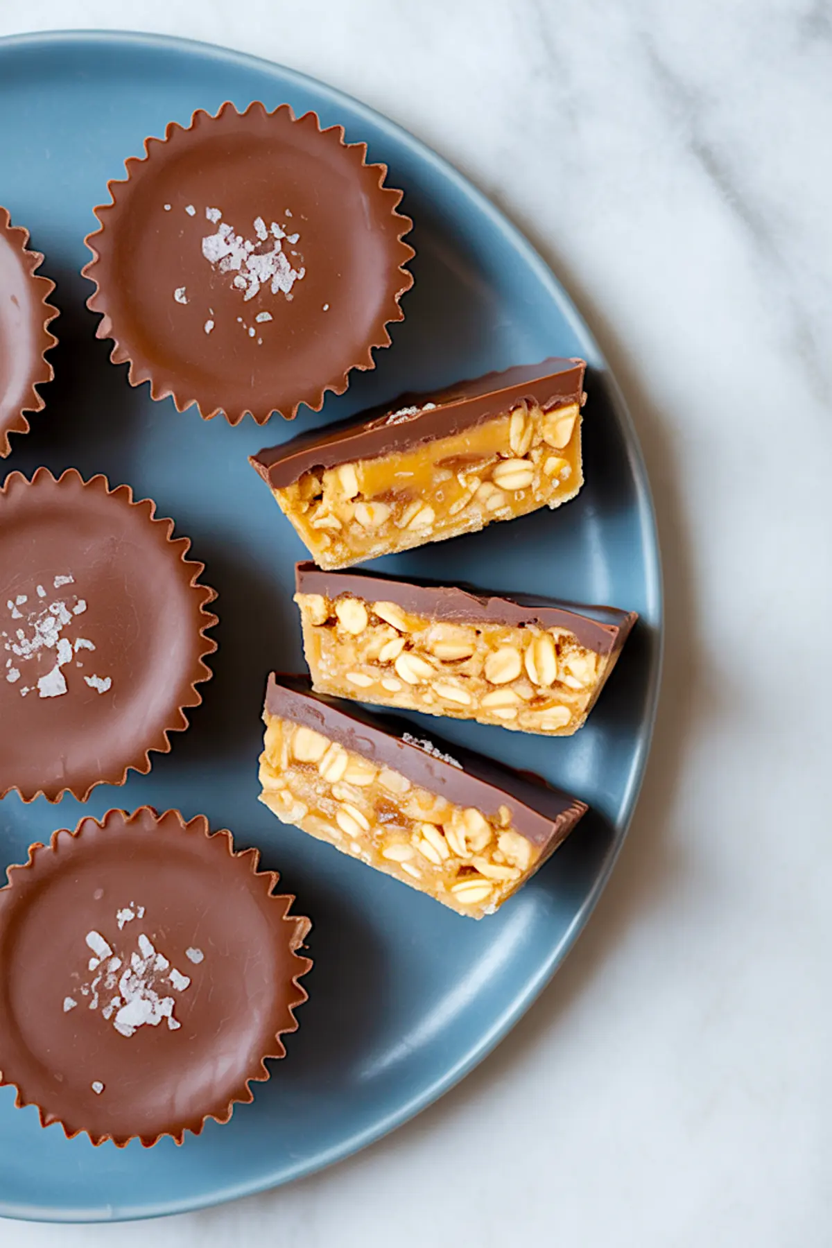 Blue plate with whole and halved no-bake oat cups, topped with sea salt flakes, revealing chewy peanut and oat filling under glossy chocolate.