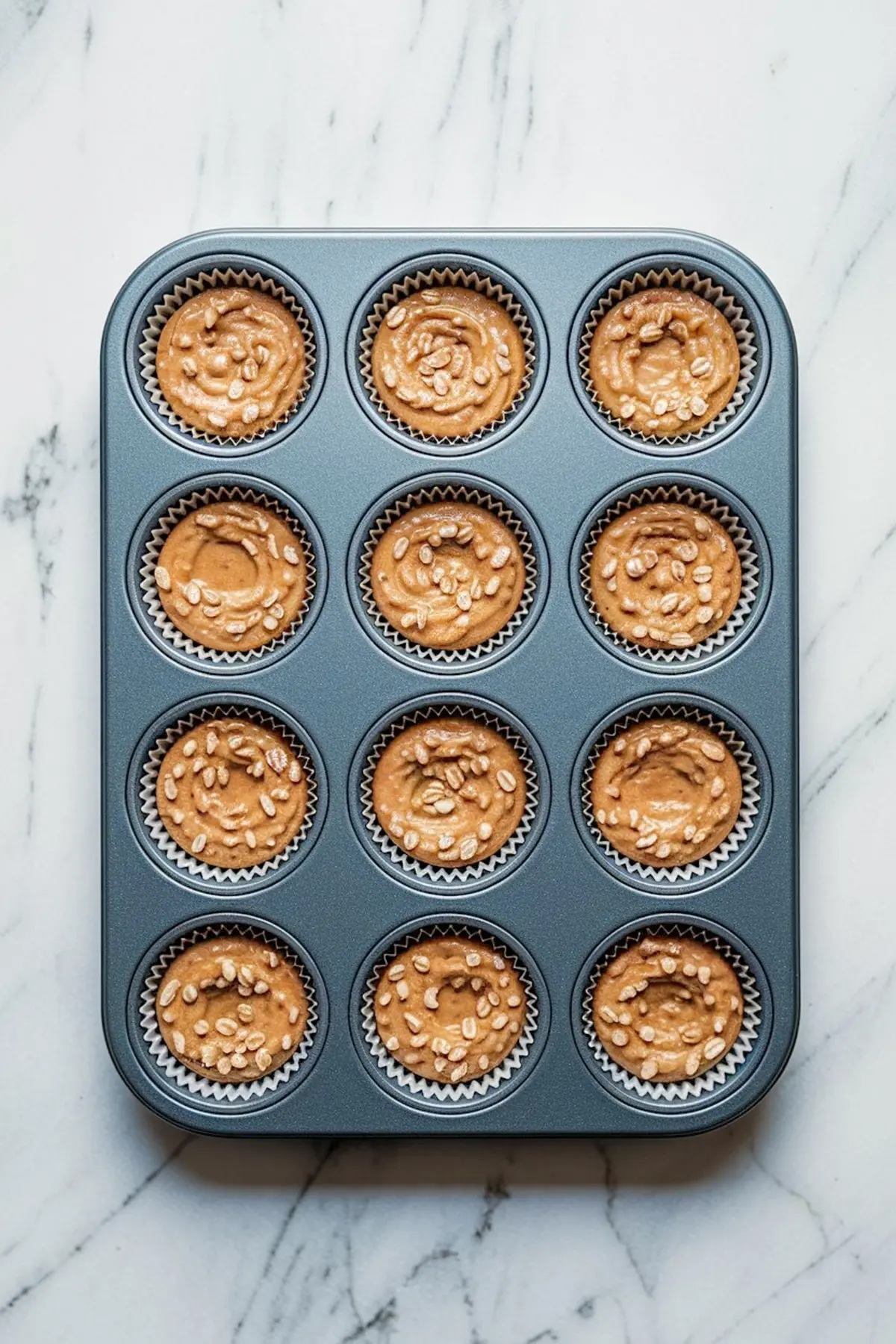 Muffin tin filled with twelve paper liners, each holding a swirled peanut butter oat batter topped with rolled oats, prepared for no-bake oat cups.