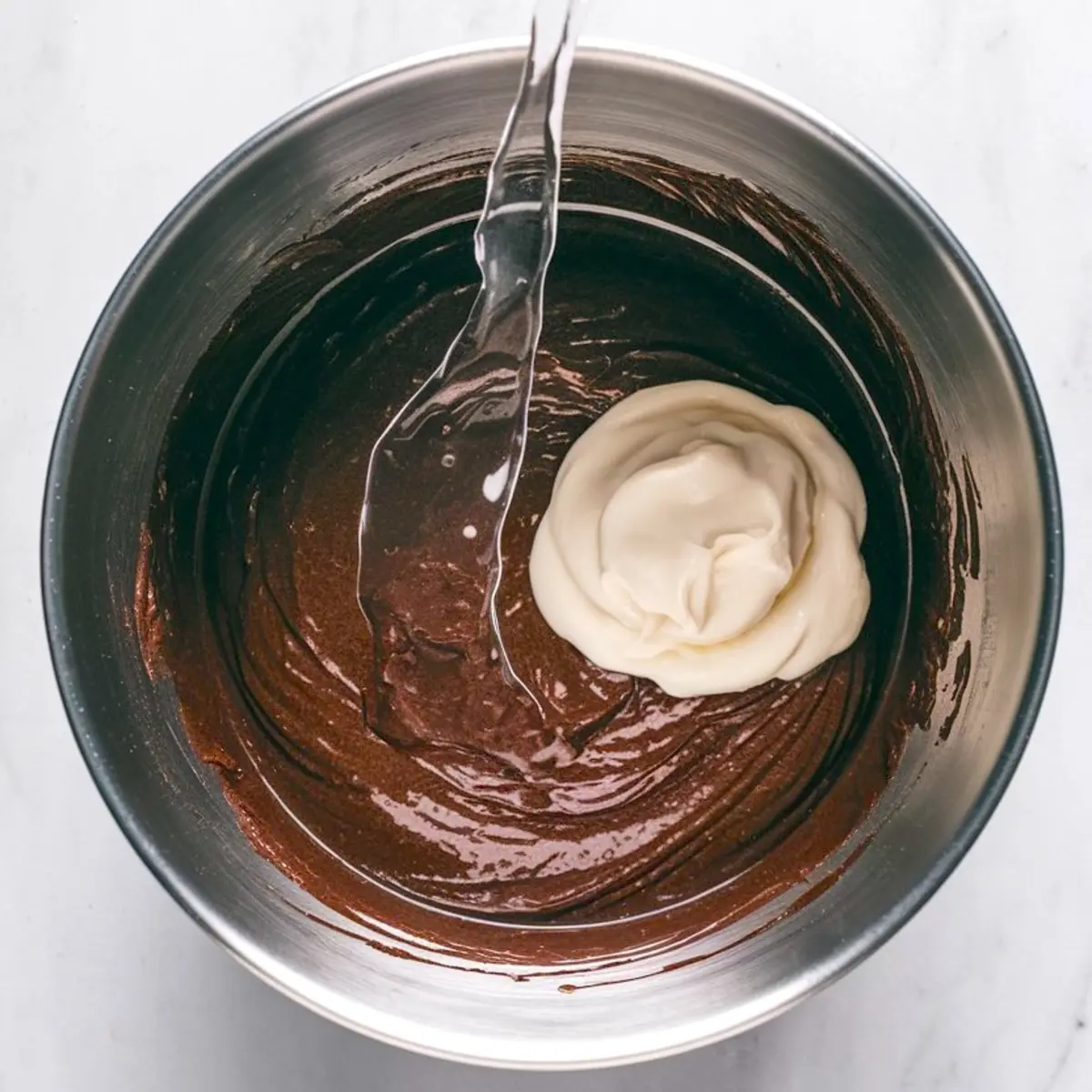 Metal mixing bowl holding partially mixed chocolate cake batter. A swirl of creamy white yogurt and a stream of water are being added to the rich chocolate mixture.