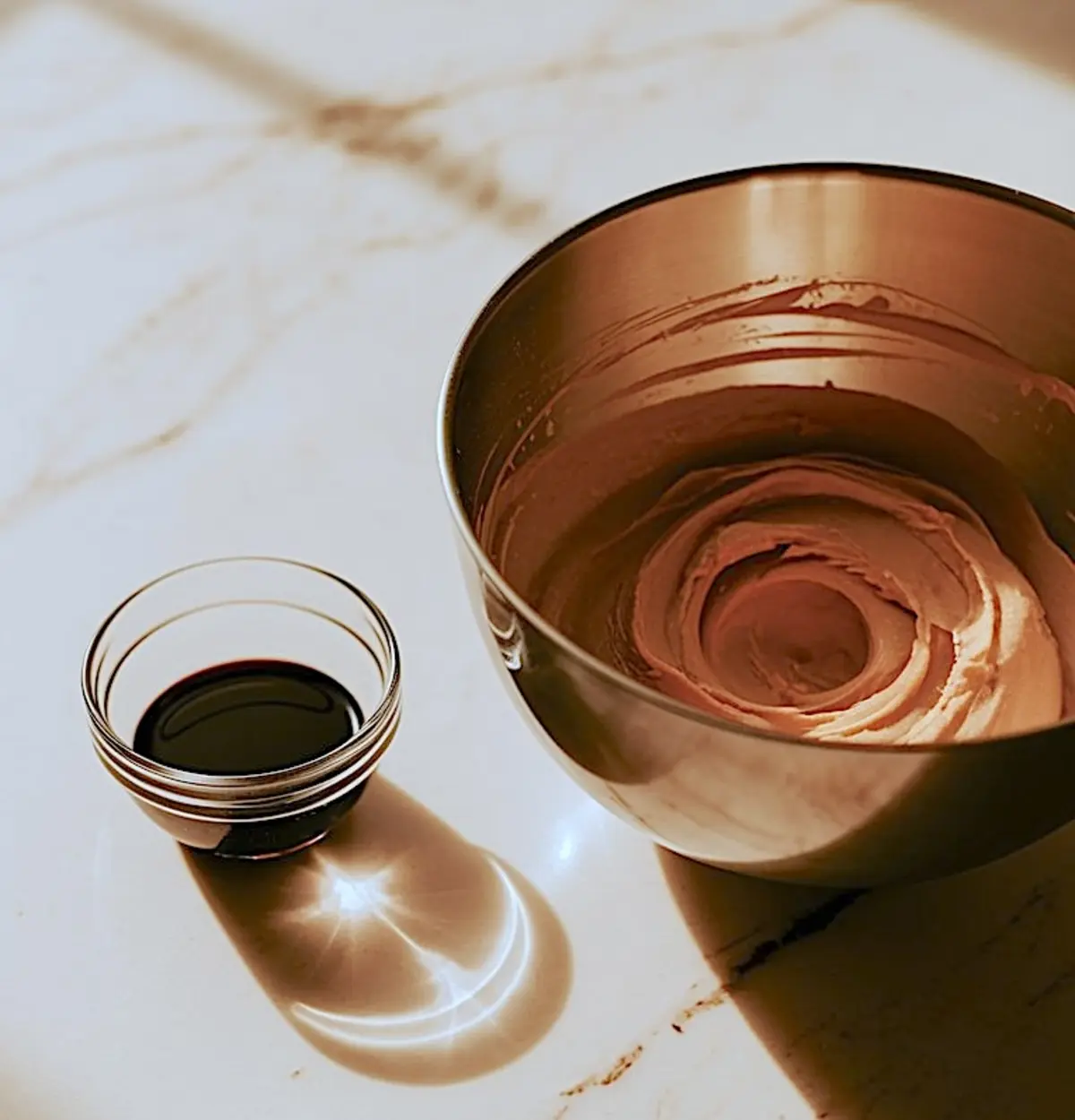Metal mixing bowl filled with creamy peanut butter icing next to a small glass bowl containing dark vanilla extract on a marble countertop, bathed in warm light.
