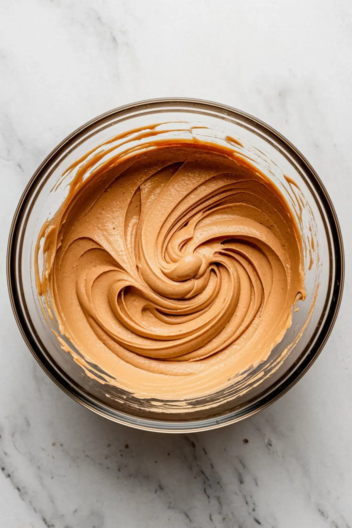 Glass bowl with smooth, freshly whipped peanut butter frosting swirled in a circular pattern on a light marble surface.
