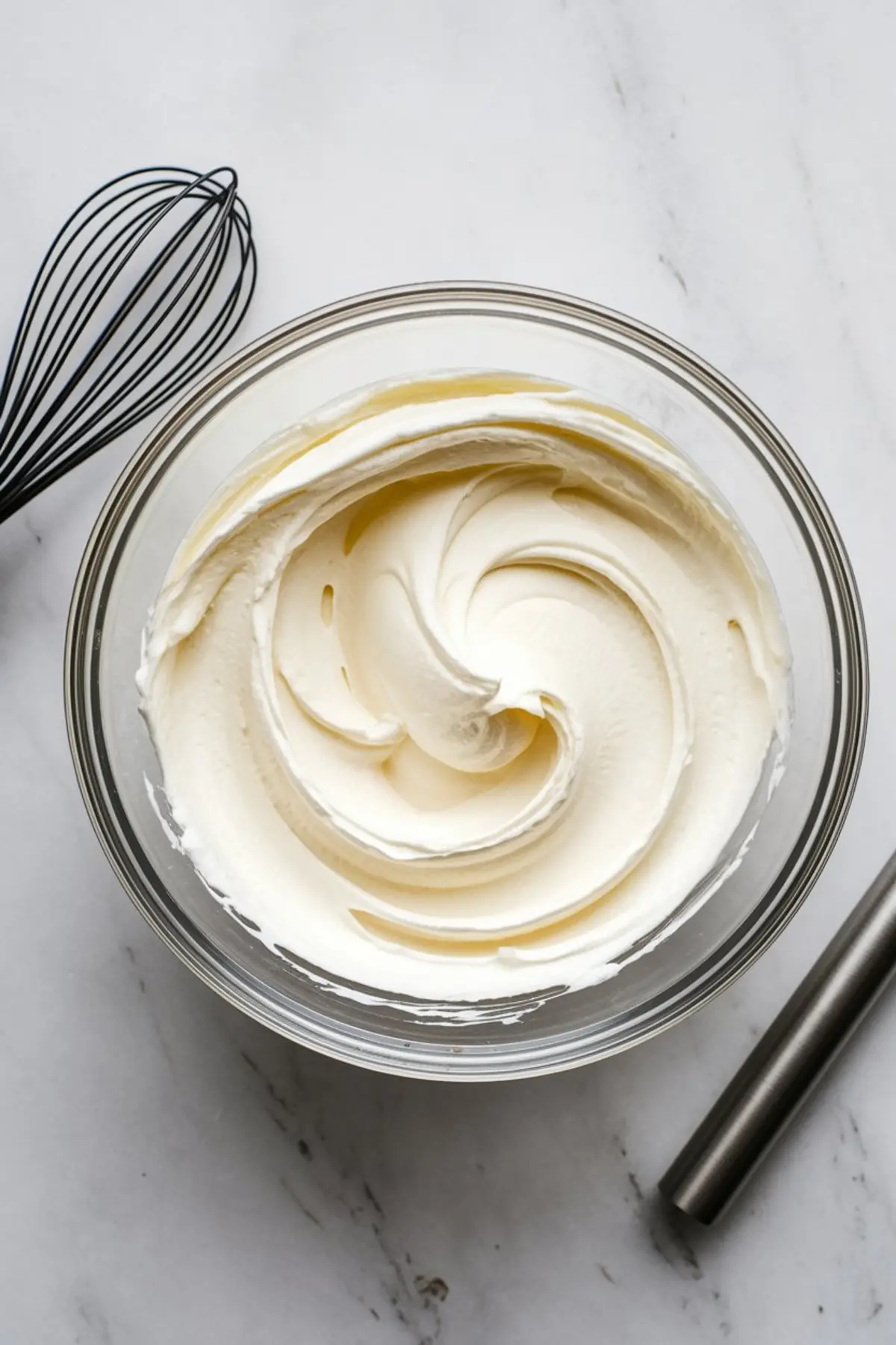 Glass bowl filled with freshly whipped cream, swirled into smooth peaks, on a marble countertop next to a metal whisk and spatula.
