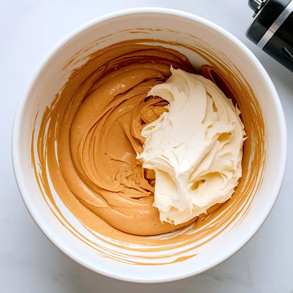 White bowl with creamy peanut butter mixture and whipped cream being blended together, with an electric mixer nearby.