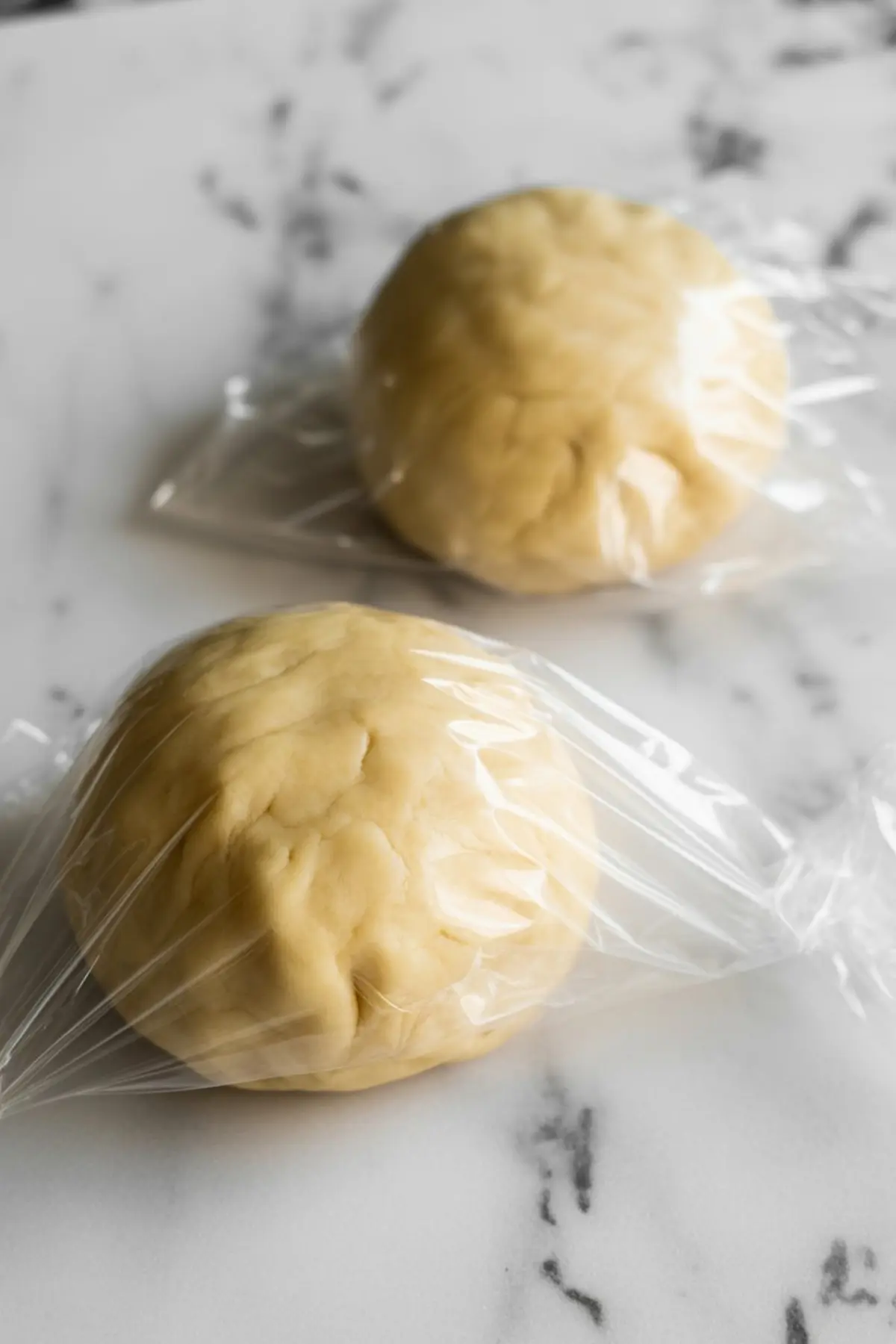 Two dough balls wrapped in plastic wrap resting on a marble countertop, prepared pie crust dough chilled and ready to roll out.
