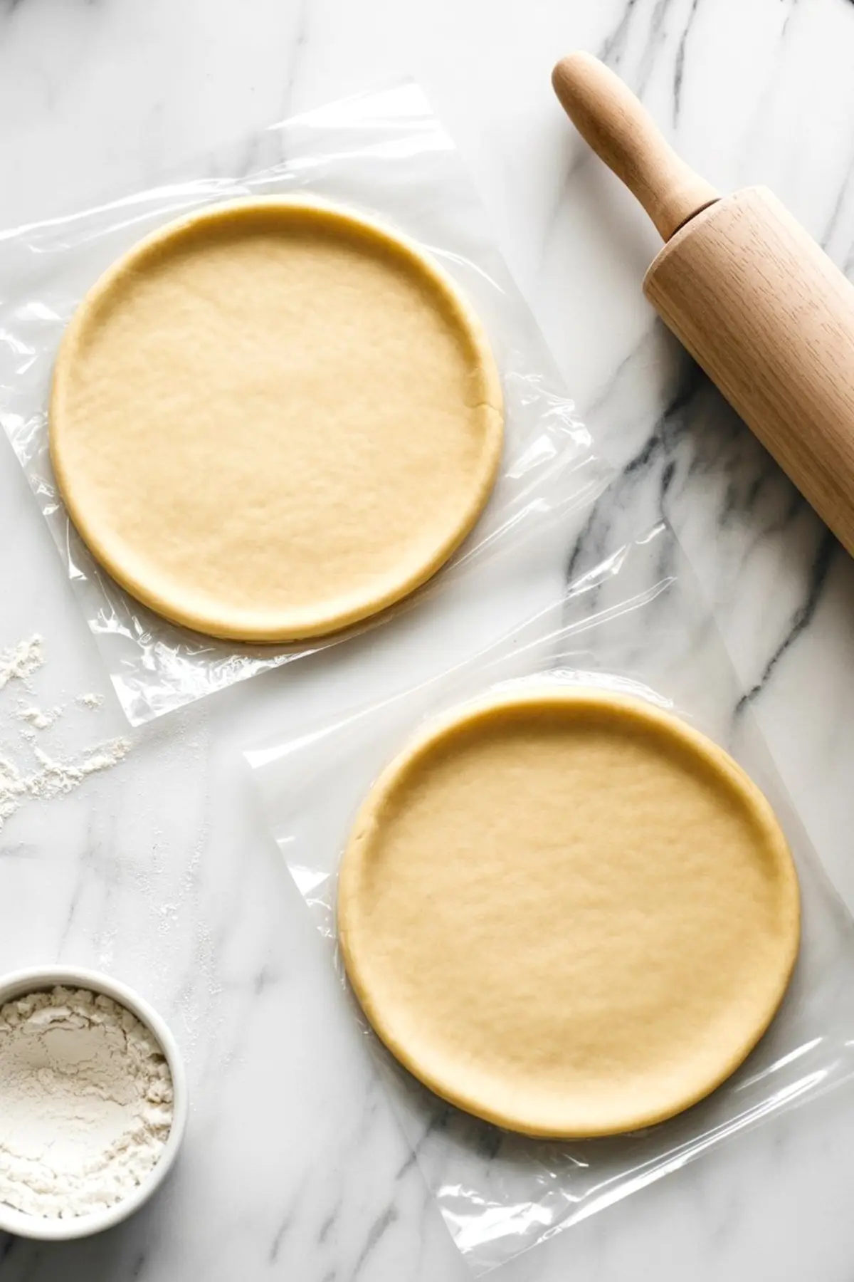 Two round, uncooked pie crusts laid on parchment paper, next to a wooden rolling pin and a small bowl of flour on a marble surface, ready for assembling homemade pineapple pie.