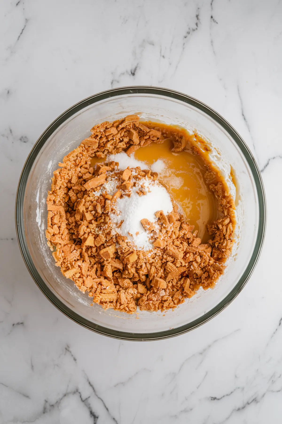 Mixing bowl containing crumbled cookies, sweetened condensed milk, and sugar on a marble surface, part of the pistachio brigadeiro base preparation.