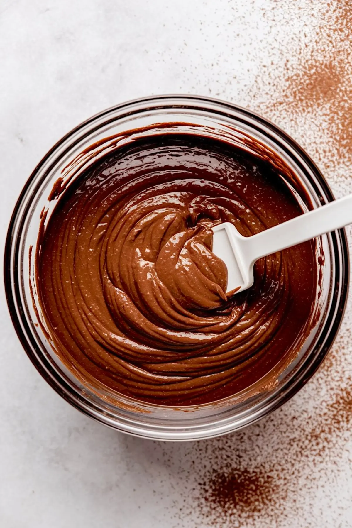 Thick chocolate batter swirled in a glass mixing bowl with a white spatula, cocoa powder dusted on the surface beneath the bowl.