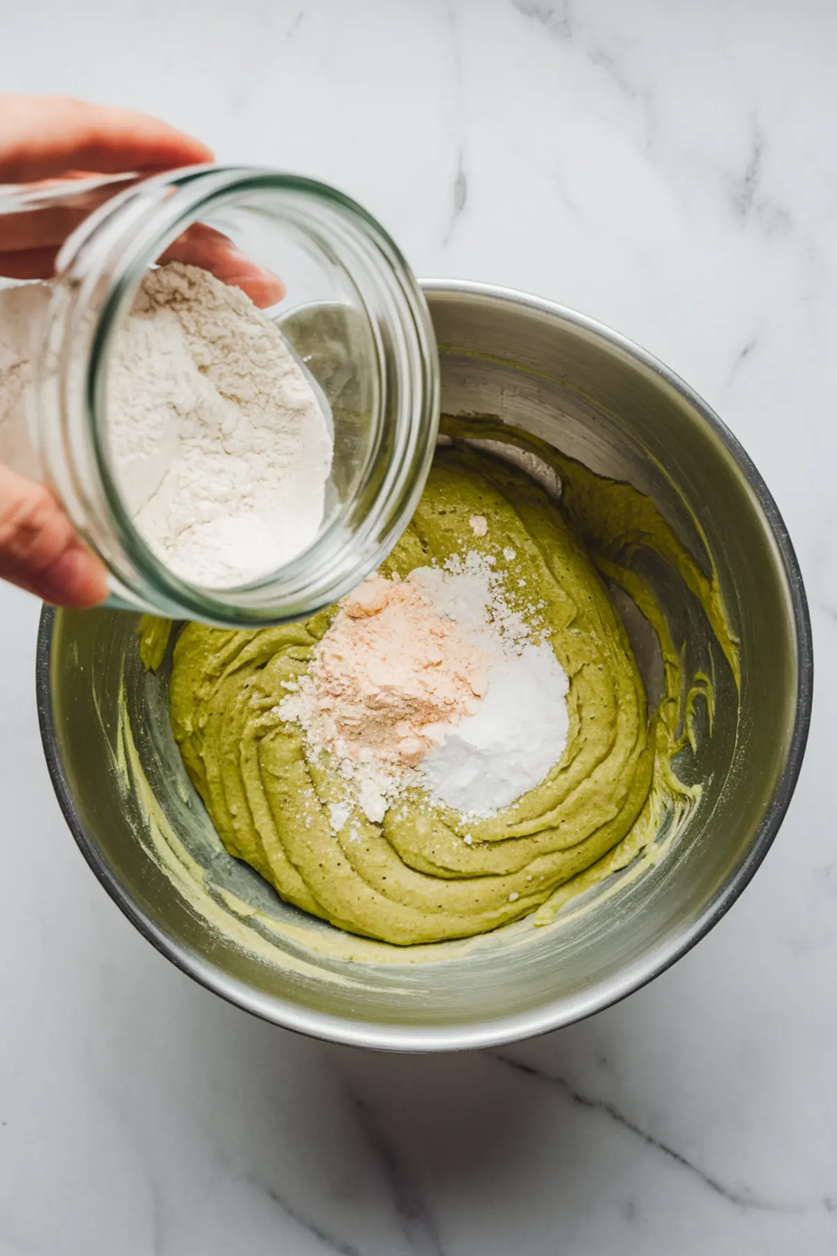Pouring flour into a mixing bowl with green pistachio cookie dough, baking powder, and baking soda on top. Preparing the batter for pistachio recipes desserts such as pistachio biscotti and pistachio bread.