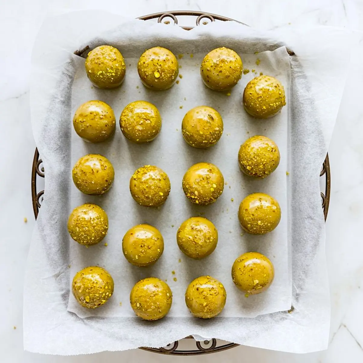 Tray lined with parchment paper holding neatly arranged pistachio ganache truffle centers, coated in a smooth green layer and sprinkled with crushed pistachios.