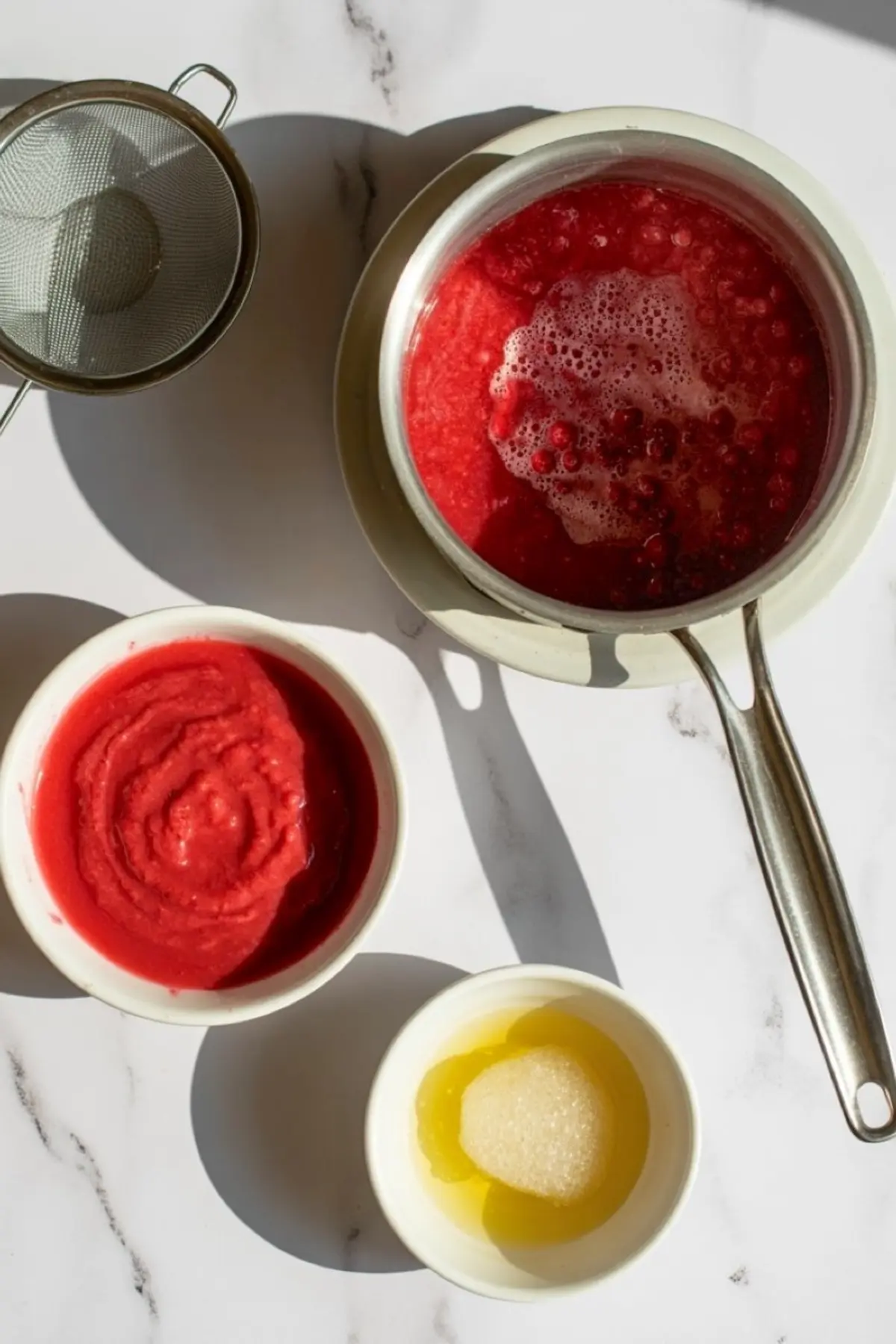 Raspberry purée being cooked in a saucepan with whole raspberries, alongside bowls containing blended raspberry mixture and hydrated gelatin, with a fine mesh strainer nearby.