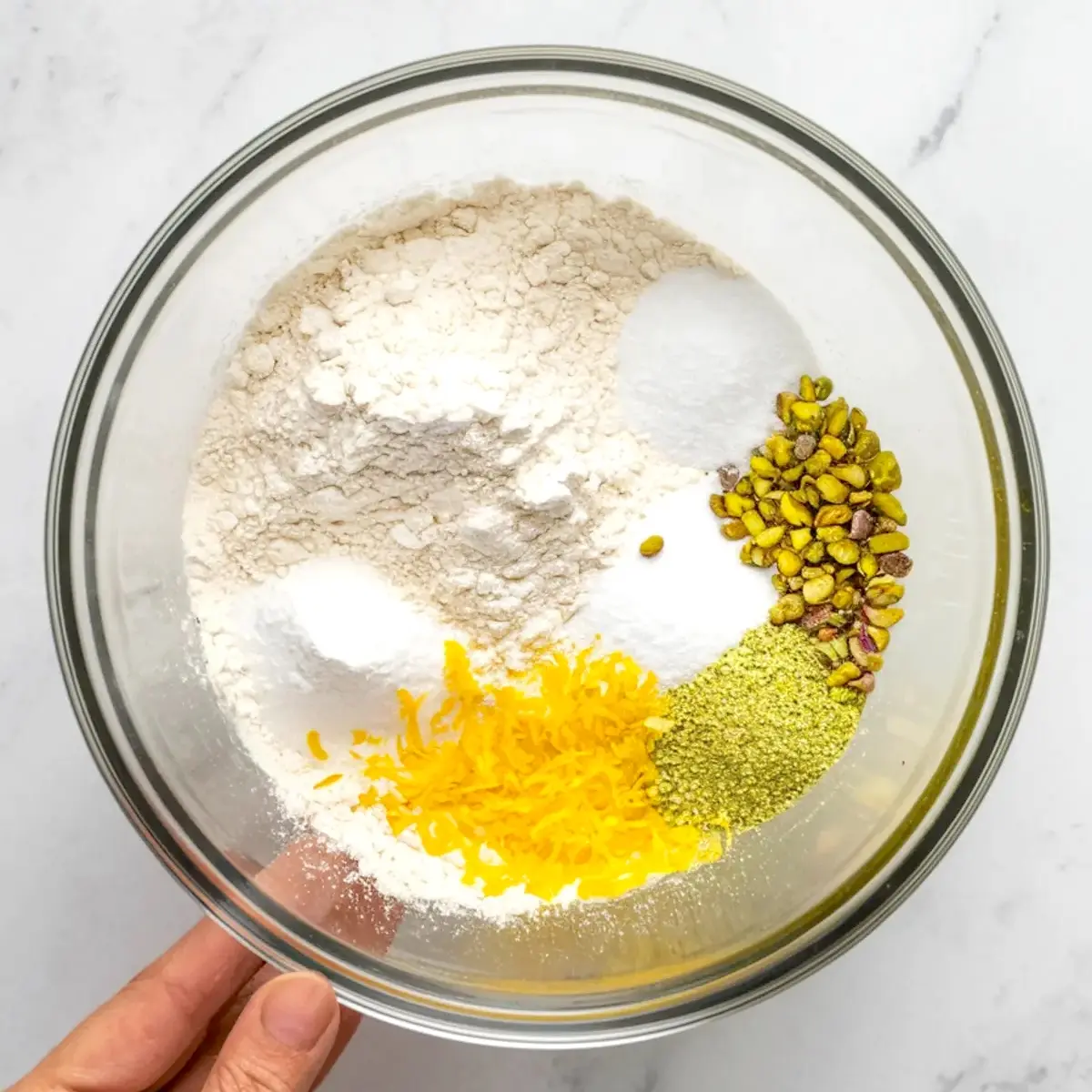 Glass bowl with separated dry ingredients including flour, baking soda, sugar, chopped pistachios, lemon zest, and pistachio powder for cookie preparation.
