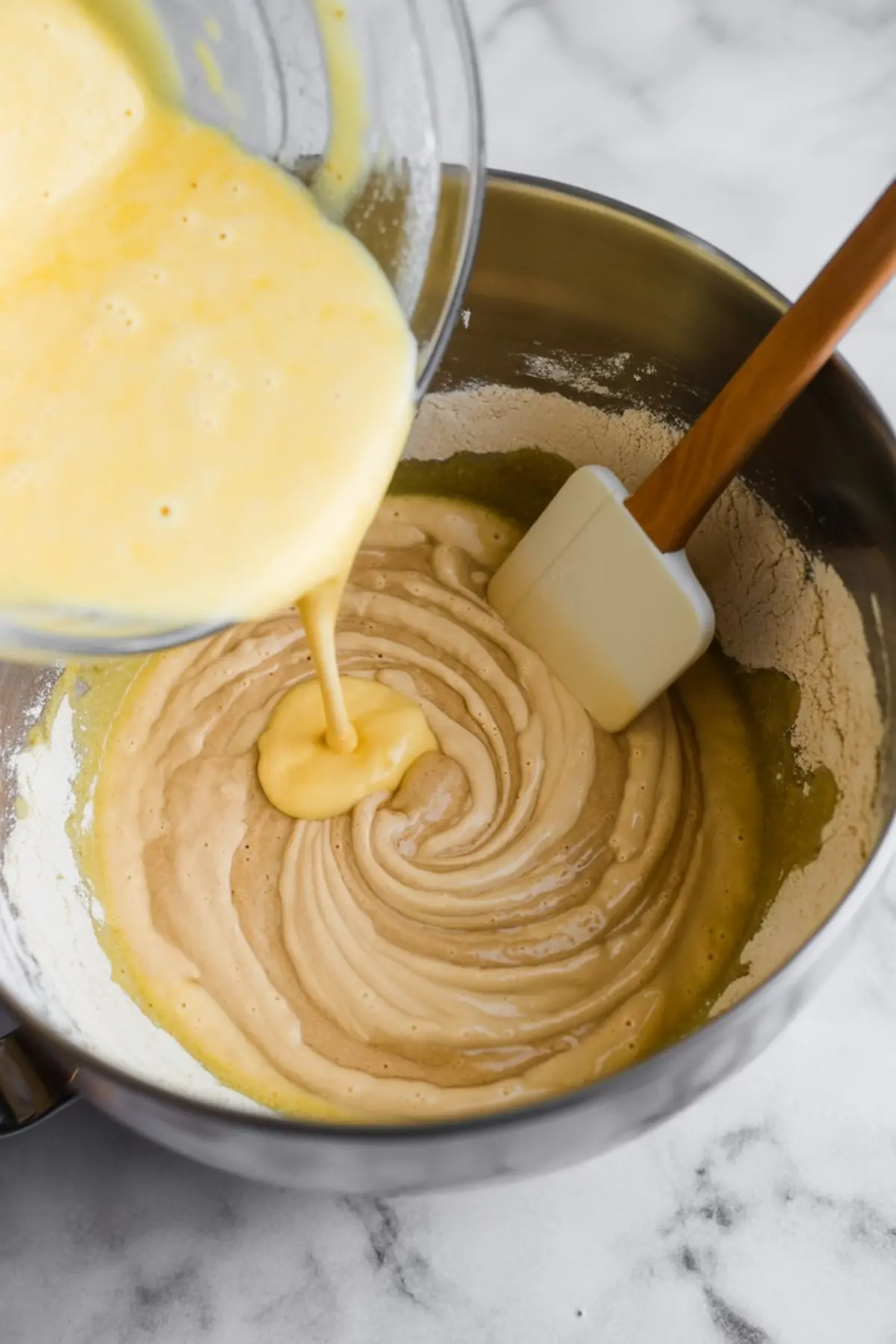 Close-up of egg mixture being poured into pumpkin batter in a metal mixing bowl with a white spatula, showing the blending of wet and dry ingredients.