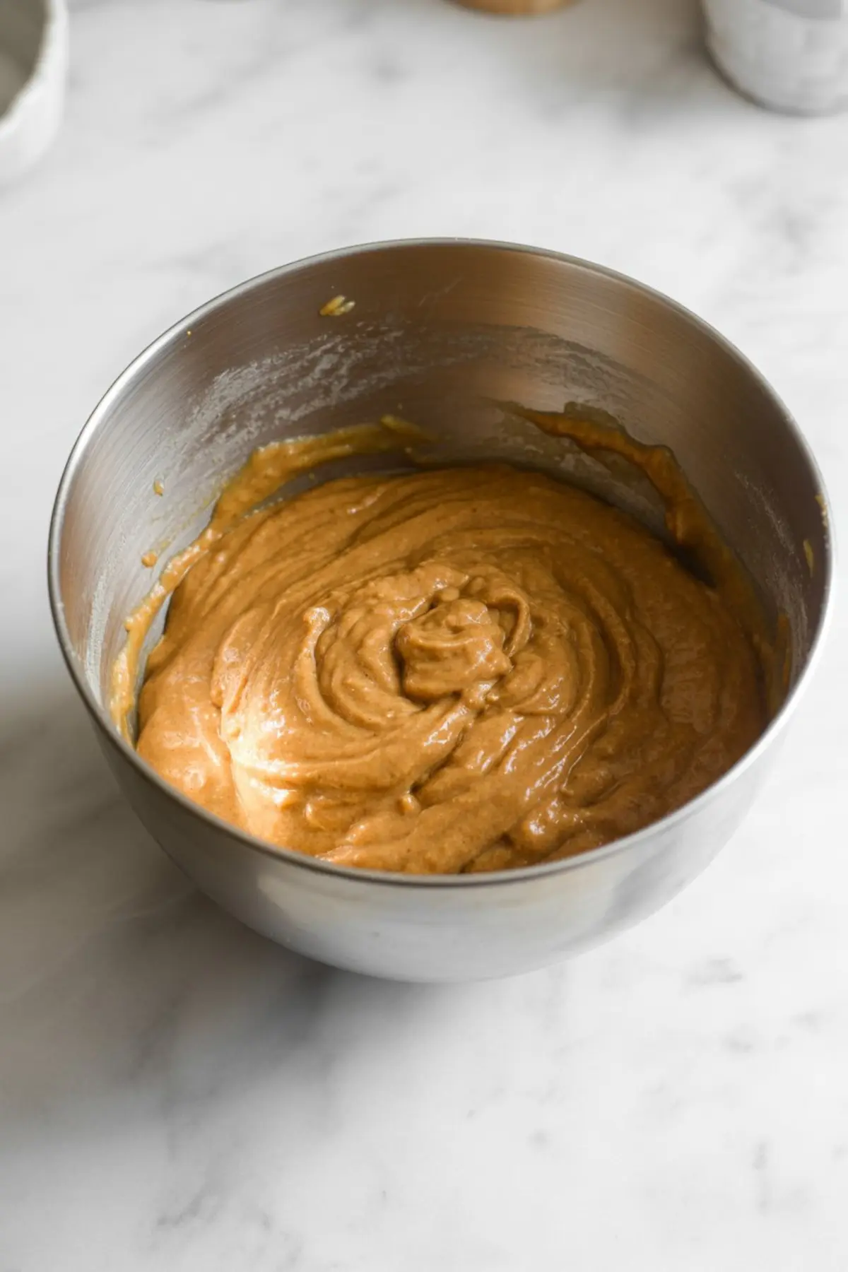 Thick pumpkin bread batter swirled in a metal mixing bowl with a creamy orange hue, ready for baking preparation.