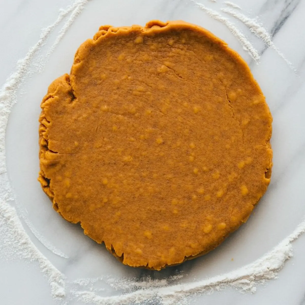 Flattened pumpkin scone dough on a floured marble surface, showing a smooth orange texture with specks of flour around the edges.