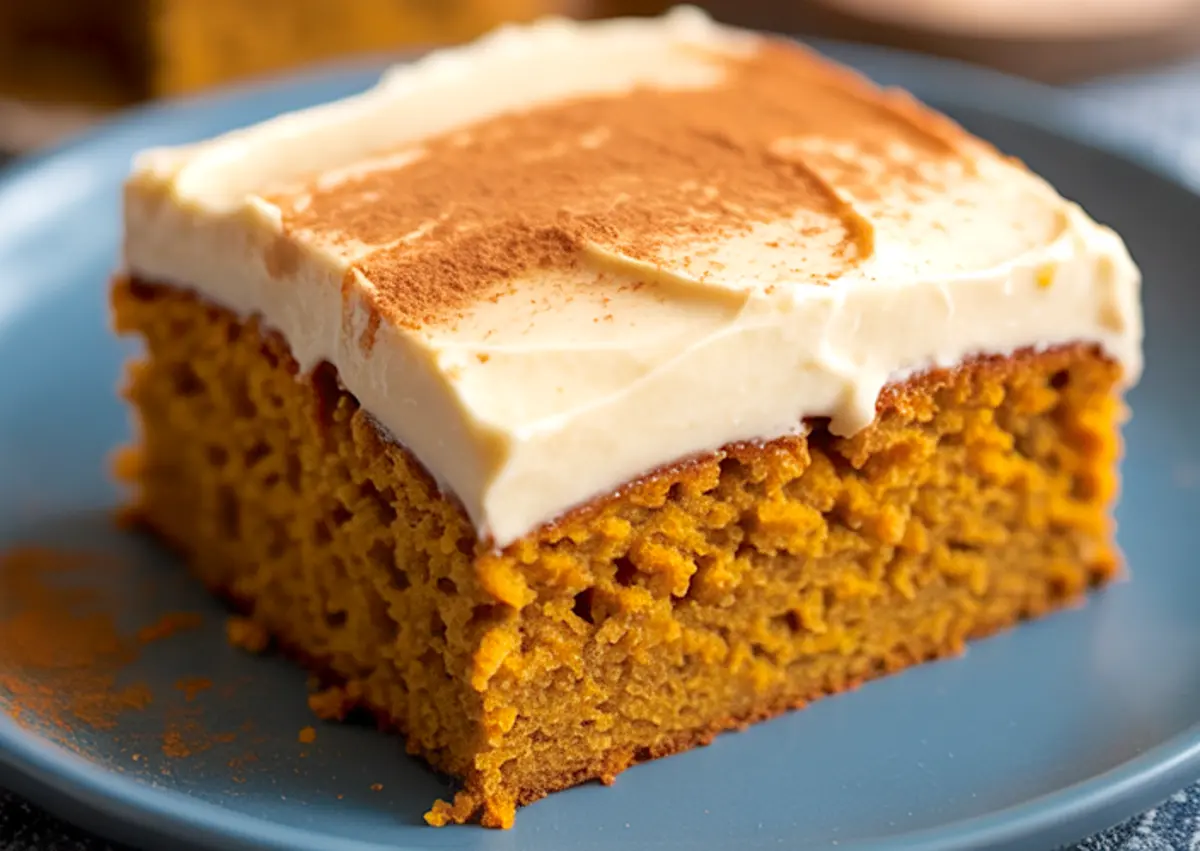 Slice of pumpkin spice cake topped with thick cream cheese frosting and a dusting of cinnamon, served on a blue plate with a moist, airy texture and golden-orange crumb.