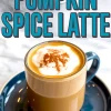 A glass mug of pumpkin spice latte topped with whipped cream and cinnamon, placed on a blue saucer with bold blue text reading “Pumpkin Spice Latte” above the mug.