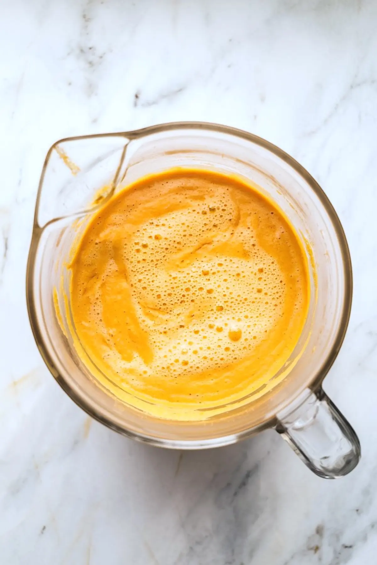A glass blender container filled with freshly blended pumpkin spice latte base, featuring a creamy, frothy orange texture on a white marble background.