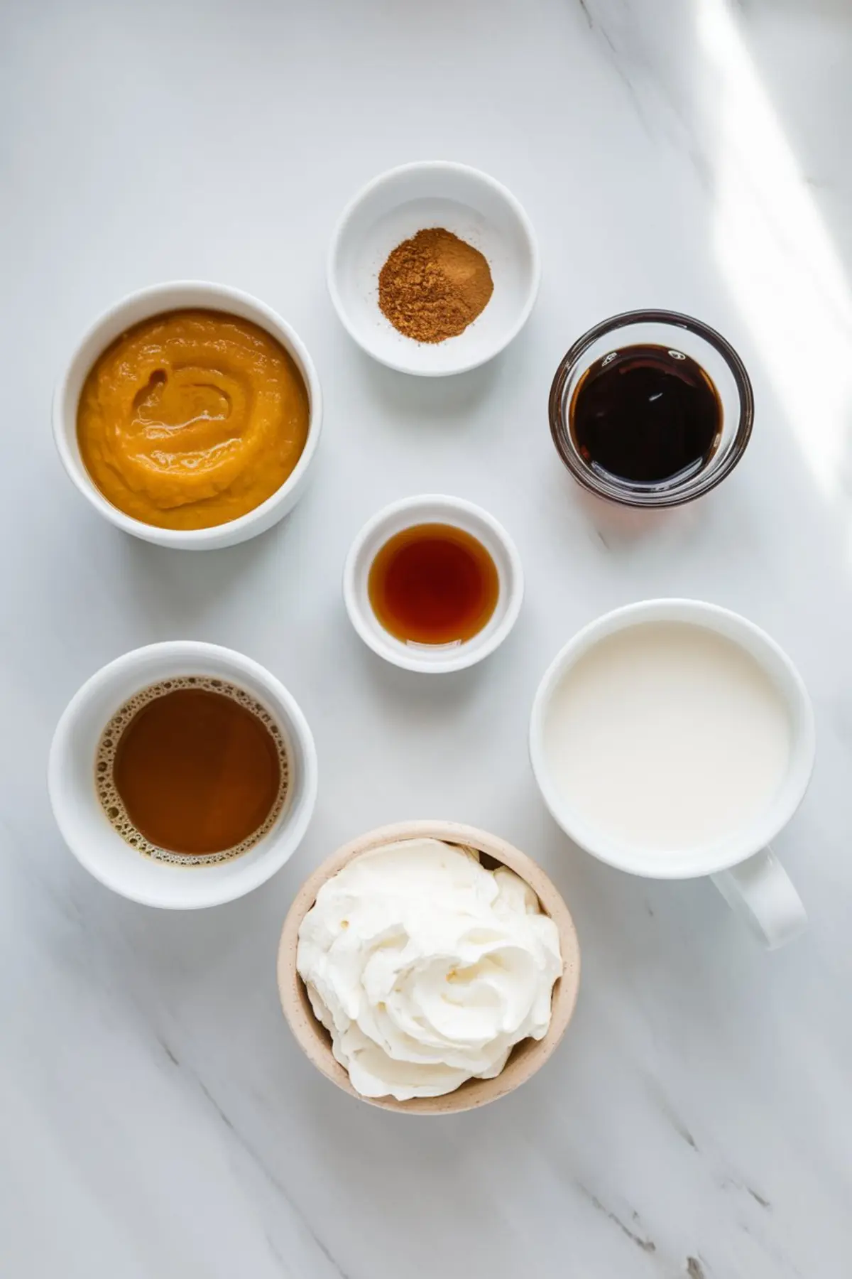 Overhead view of pumpkin spice latte ingredients in individual bowls, including pumpkin puree, cinnamon, espresso, vanilla extract, maple syrup, milk, and whipped cream on a white marble surface.