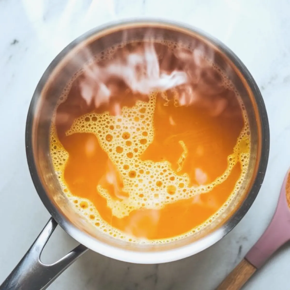 A steaming saucepan of pumpkin spice latte mixture with visible froth and swirling texture, captured mid-simmer on a marble countertop.