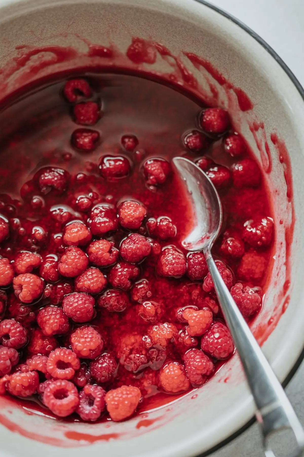 Close-up of fresh raspberries simmering in a red sauce inside a mixing bowl, with a silver spoon partially submerged in the glossy raspberry filling.