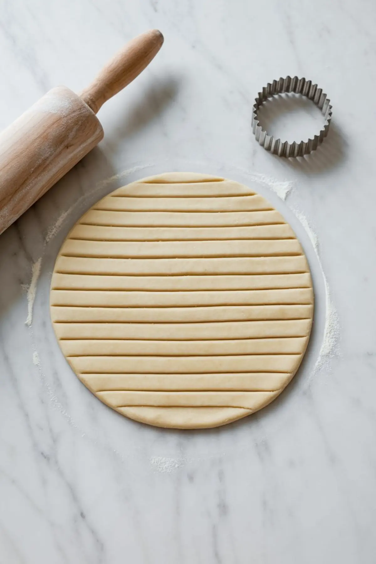 Rolled-out pie crust cut into narrow lattice strips, placed on a floured marble surface with a fluted biscuit cutter and rolling pin.