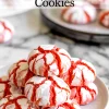 A plate of strawberry crinkle cookies stacked in a pyramid, featuring vibrant red cracks and a white powdered sugar coating, set on a marble surface with additional cookies blurred in the background.