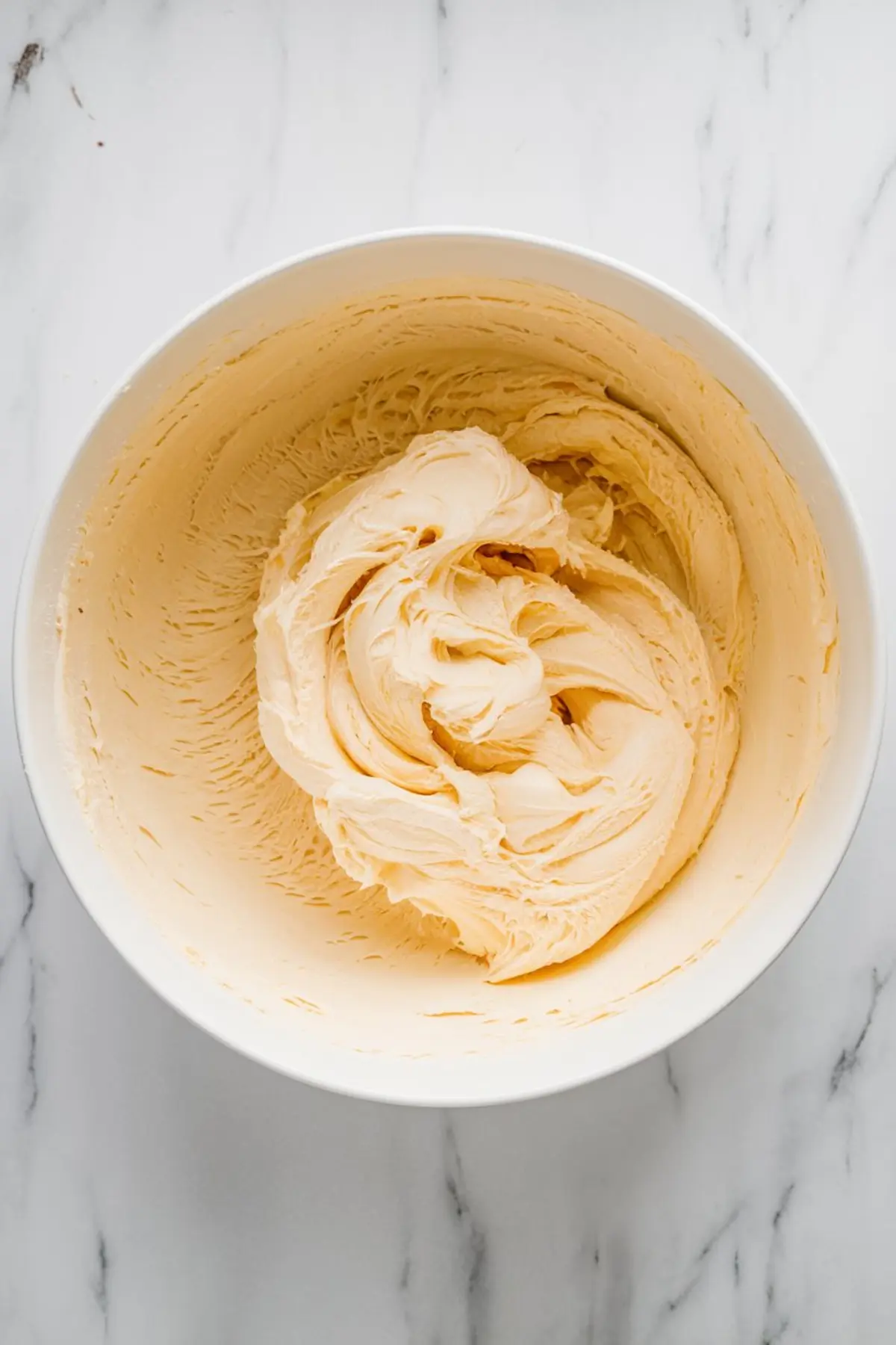 A mixing bowl filled with smooth, pale yellow buttercream frosting on a marble countertop, ideal for cookie or cake decorating recipes.