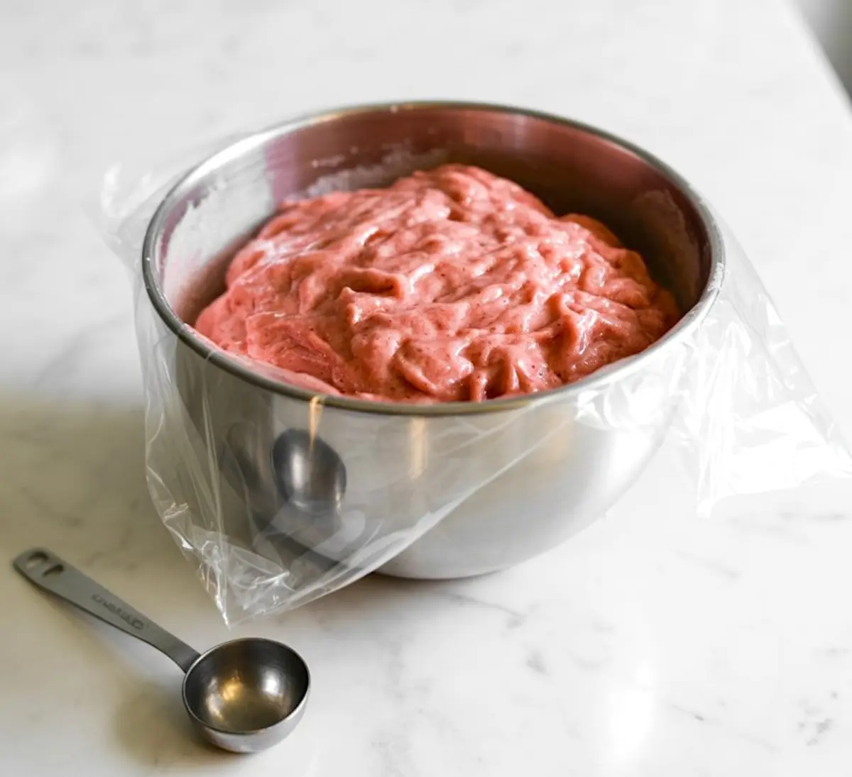 Mixing bowl filled with pink strawberry cookie dough, partially covered with plastic wrap and placed on a marble counter with a metal tablespoon beside it.
