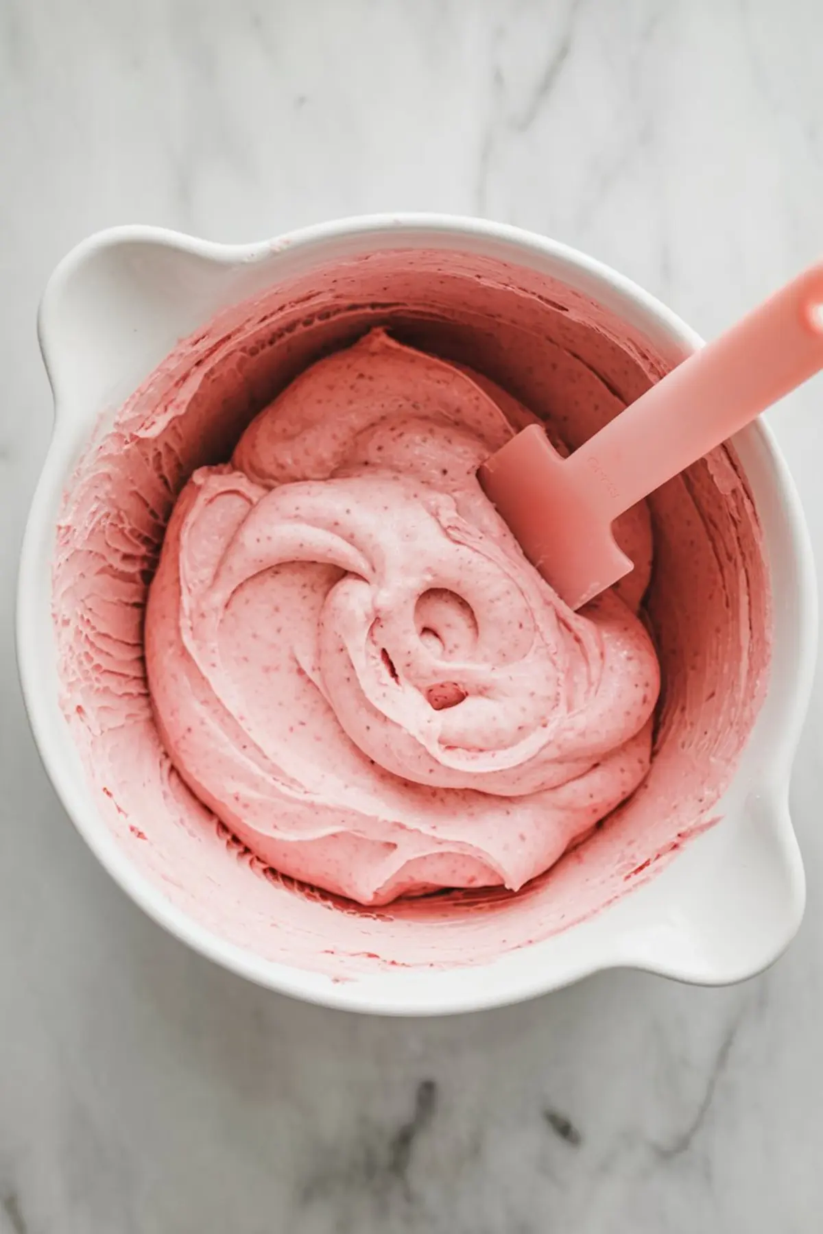 A white mixing bowl filled with fluffy pink strawberry frosting, with a pink silicone spatula resting inside, ideal for decorating desserts.