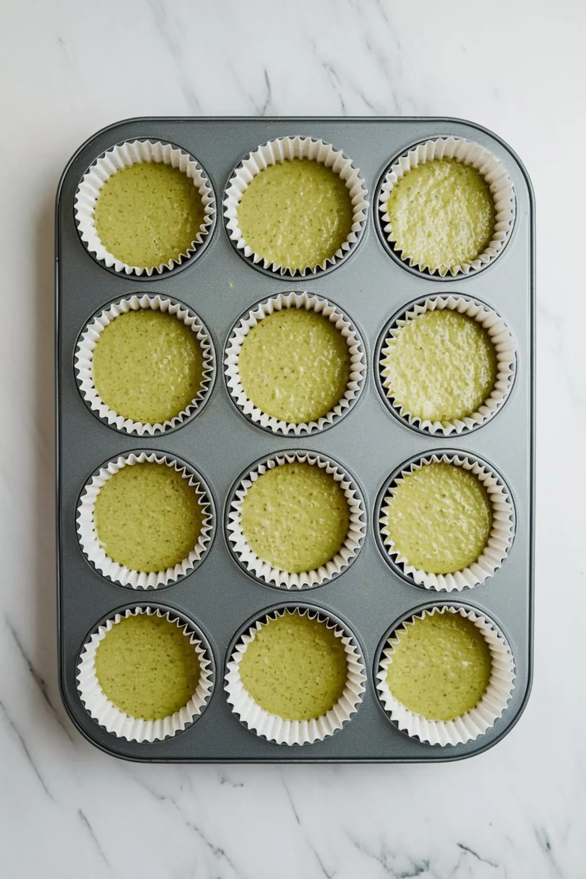 A metal muffin pan holding twelve paper liners filled with raw matcha cupcake batter, positioned on a white marble surface, ready for baking.