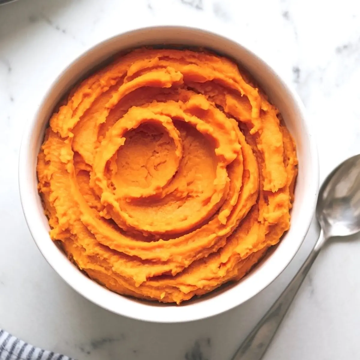 A white bowl filled with creamy mashed sweet potatoes swirled in a spiral pattern, placed next to a metal spoon on a marble surface.