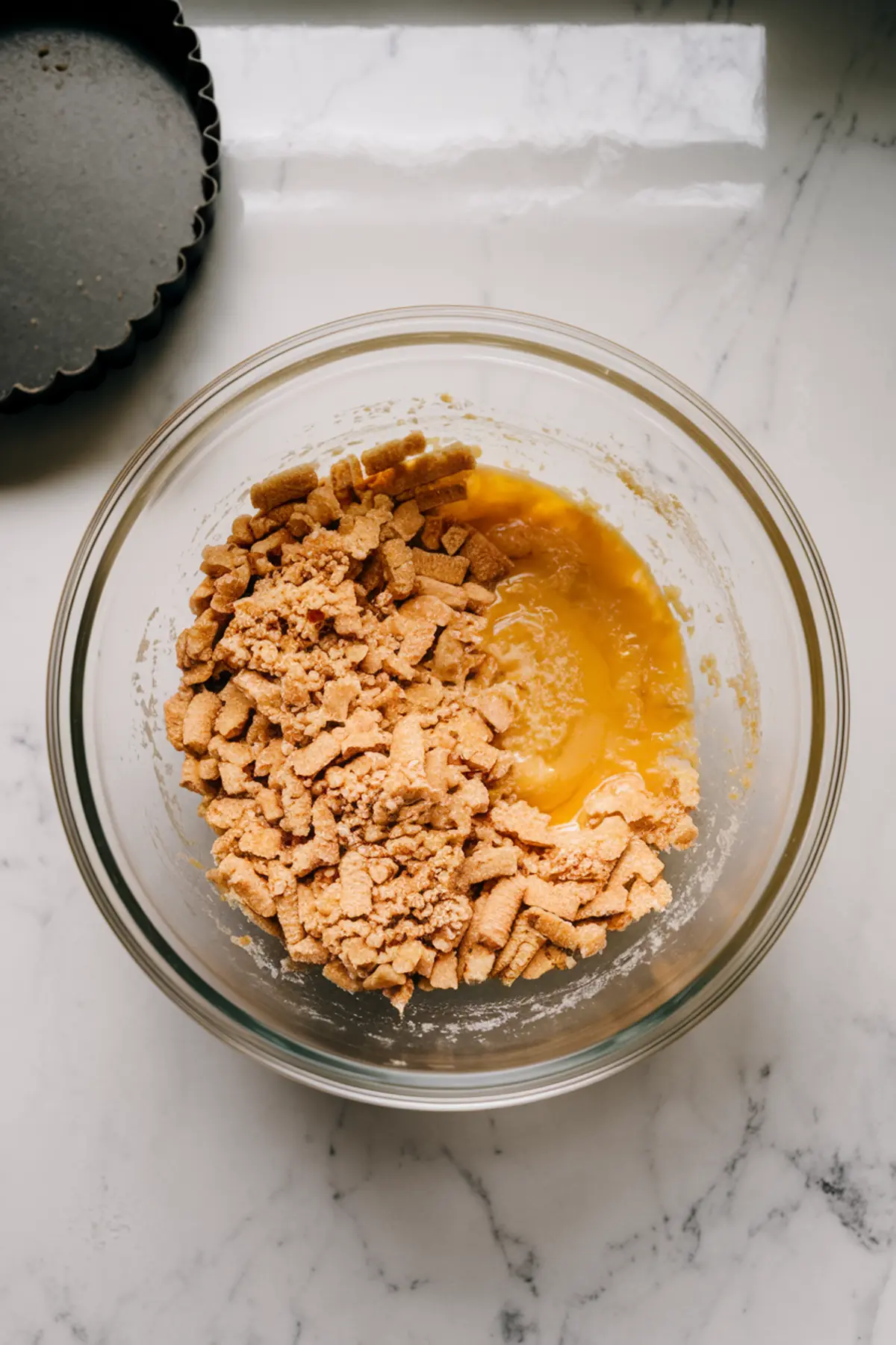 Crushed biscuits mixed with melted butter in a glass mixing bowl, captured before forming the crust for a pie or cheesecake.