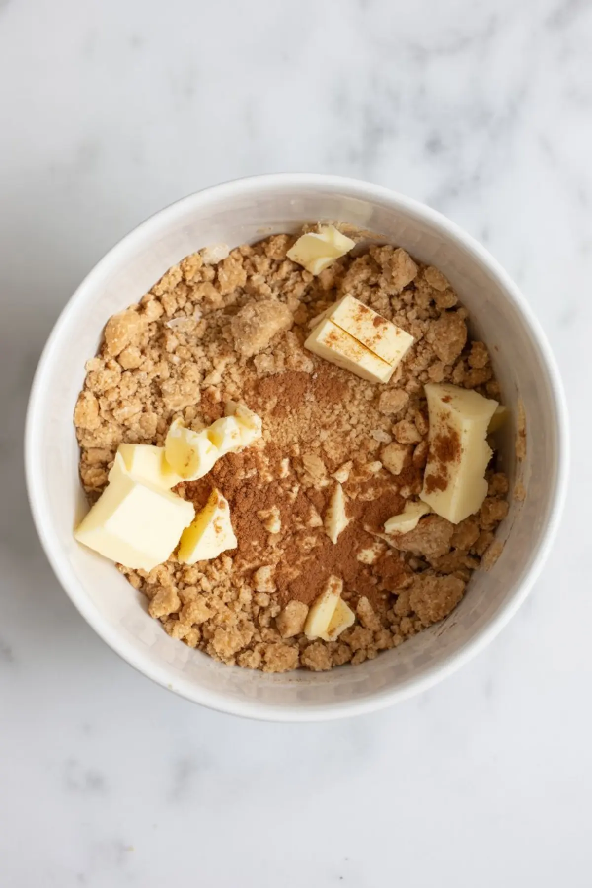 White bowl filled with oat crumble topping ingredients, including cubed butter and cinnamon over a brown sugar and flour mixture.